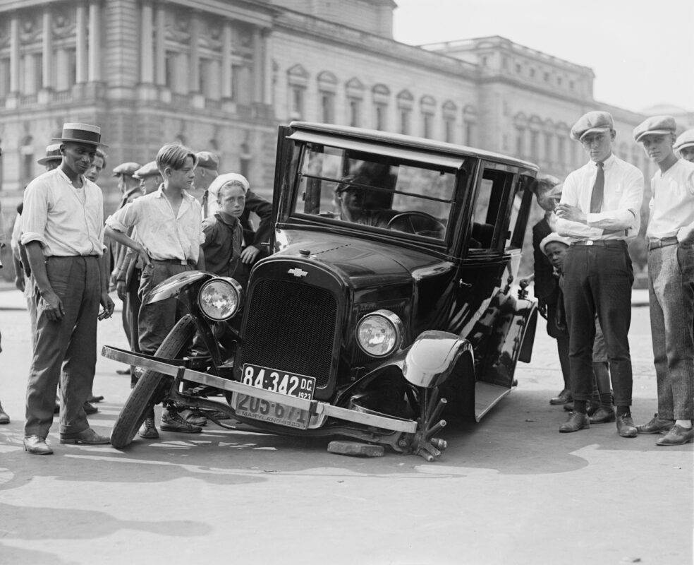 A vintage car crash attended by a group of onlookers in a 1920s urban setting.